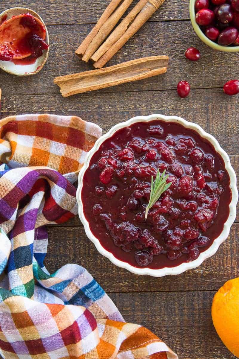 Overhead portrait image of a light-colored bowl filled with Boozy Spiced Cranberry Sauce surrounded by the rest of the ingredients in the recipe.