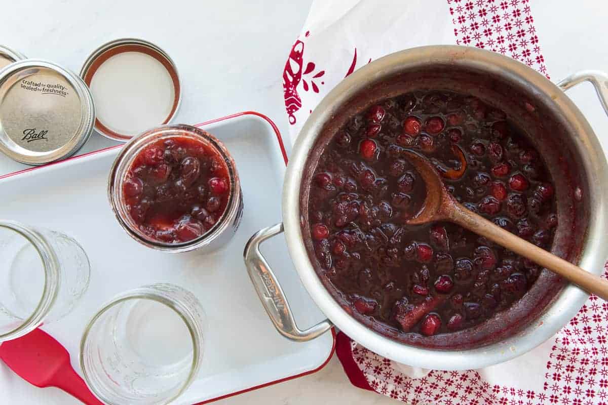 Overhead image of a silver pot with Boozy Spiced Cranberry Sauce that has been spooned into a glass mason jar on the right.