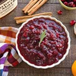 Preview image of the Boozy Spiced Cranberry Sauce in a light-colored bowl. The ingredients surround the bowl on a dark wooden surface.