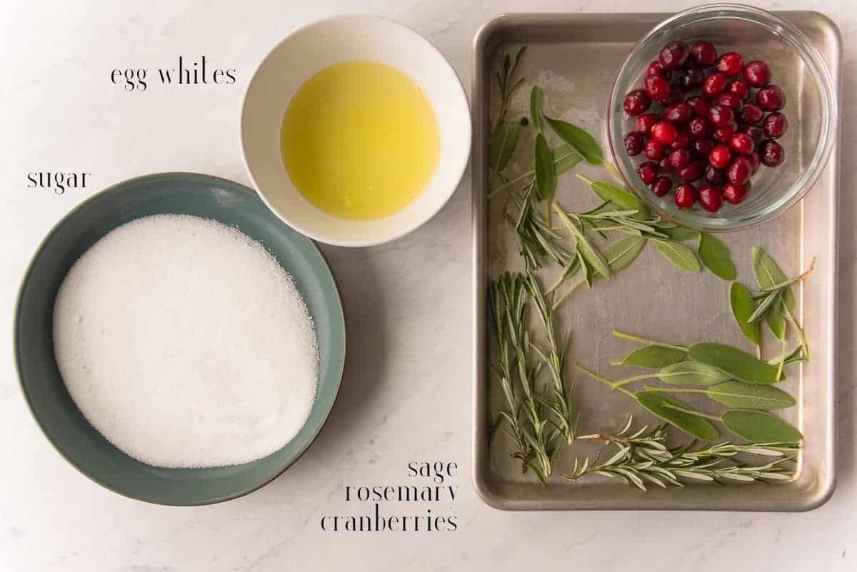 The ingredients to make sugared cranberries and fresh herbs: egg whites, fresh cranberries, sage, rosemary, and sugar