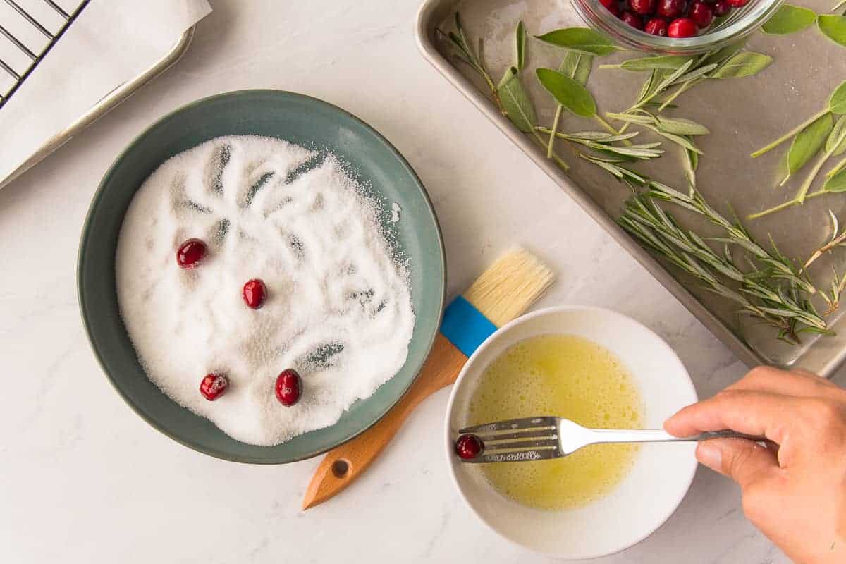 A hand uses a fork to dip the cranberries in egg whites before tossing them in a green bowl of granulated sugar.