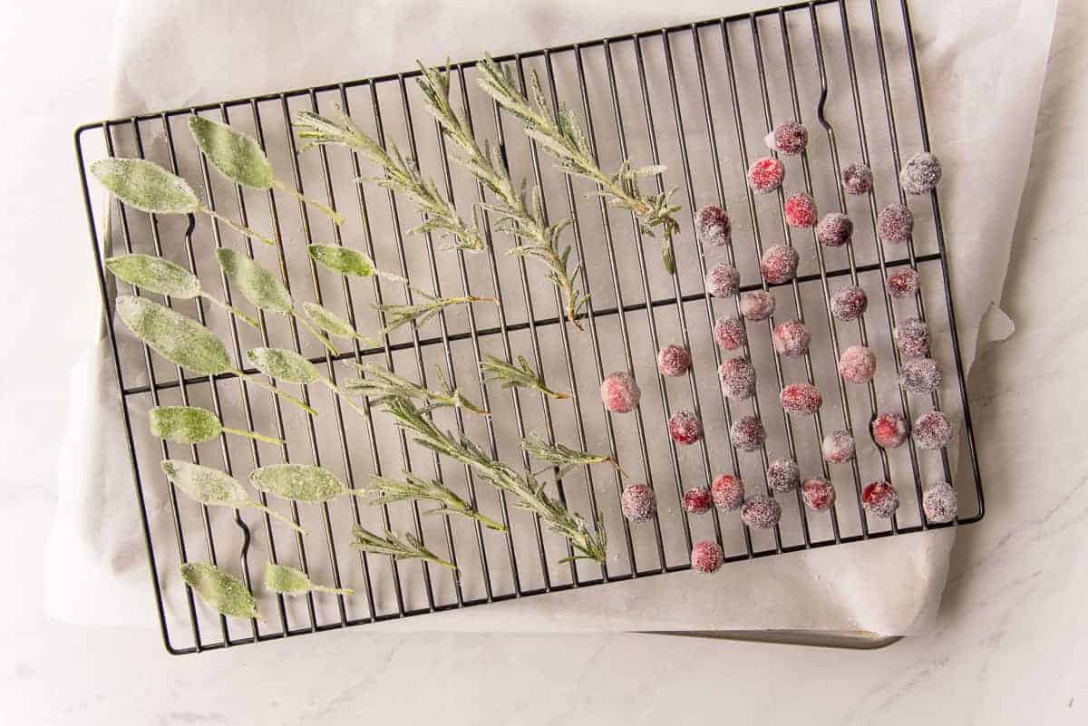 The sugared cranberries, sage, and rosemary dry on a black cooling rack set atop a parchment-lined sheetpan.