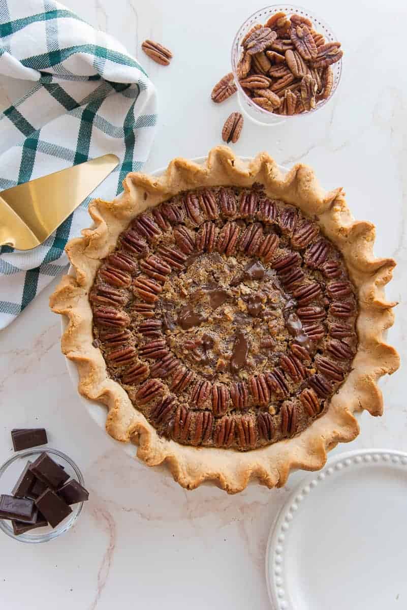 Overhead portrait image of the baked Maple Bourbon Pecan Pie with Chocolate Chunks next to a plaid green and white kitchen towel, a bowl of pecans, and bowl of chocolate chunks.