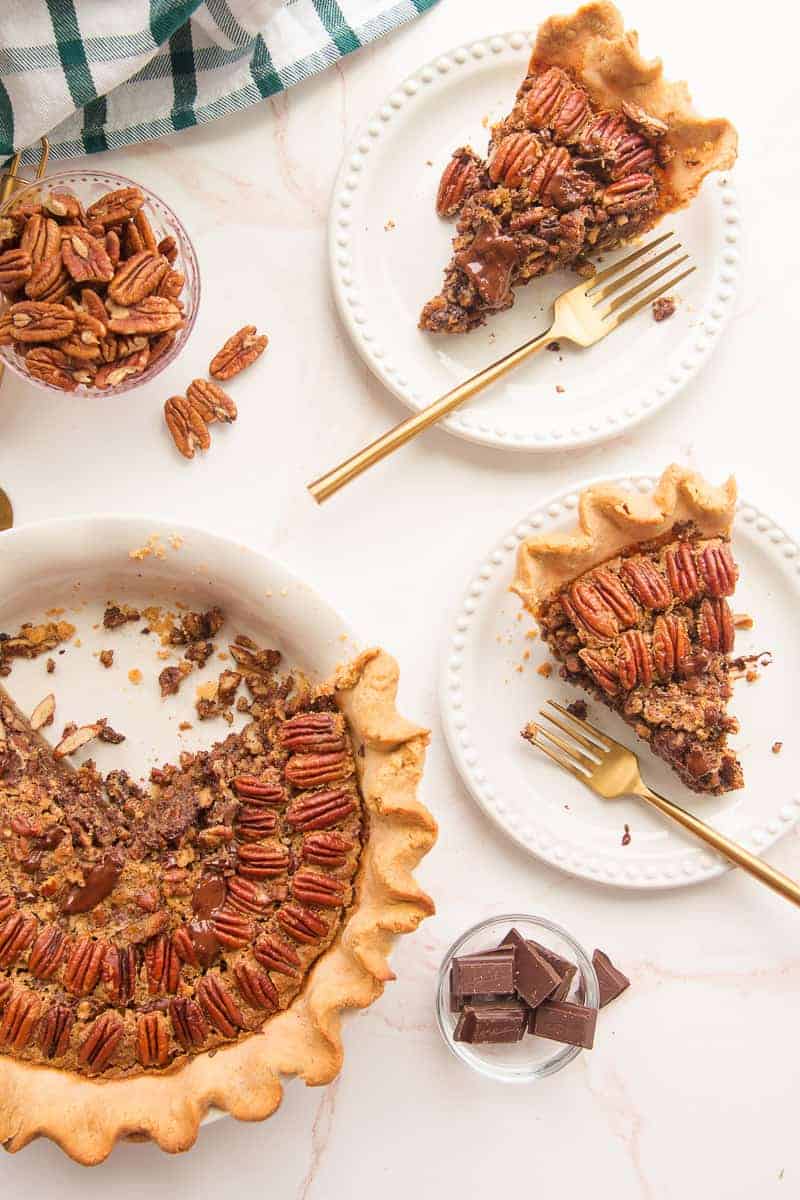 Overhead image of the cut Maple Bourbon Pecan Pie with Chocolate Chunks next to two white plates with slices of the pie and gold forks on them.