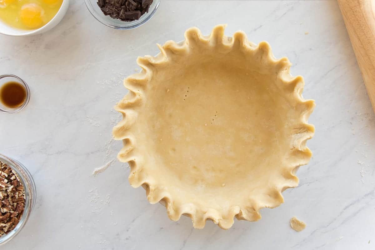 The mealy pie dough shell is pictured next to a rolling pin, chocolate, eggs, vanilla, and nuts.