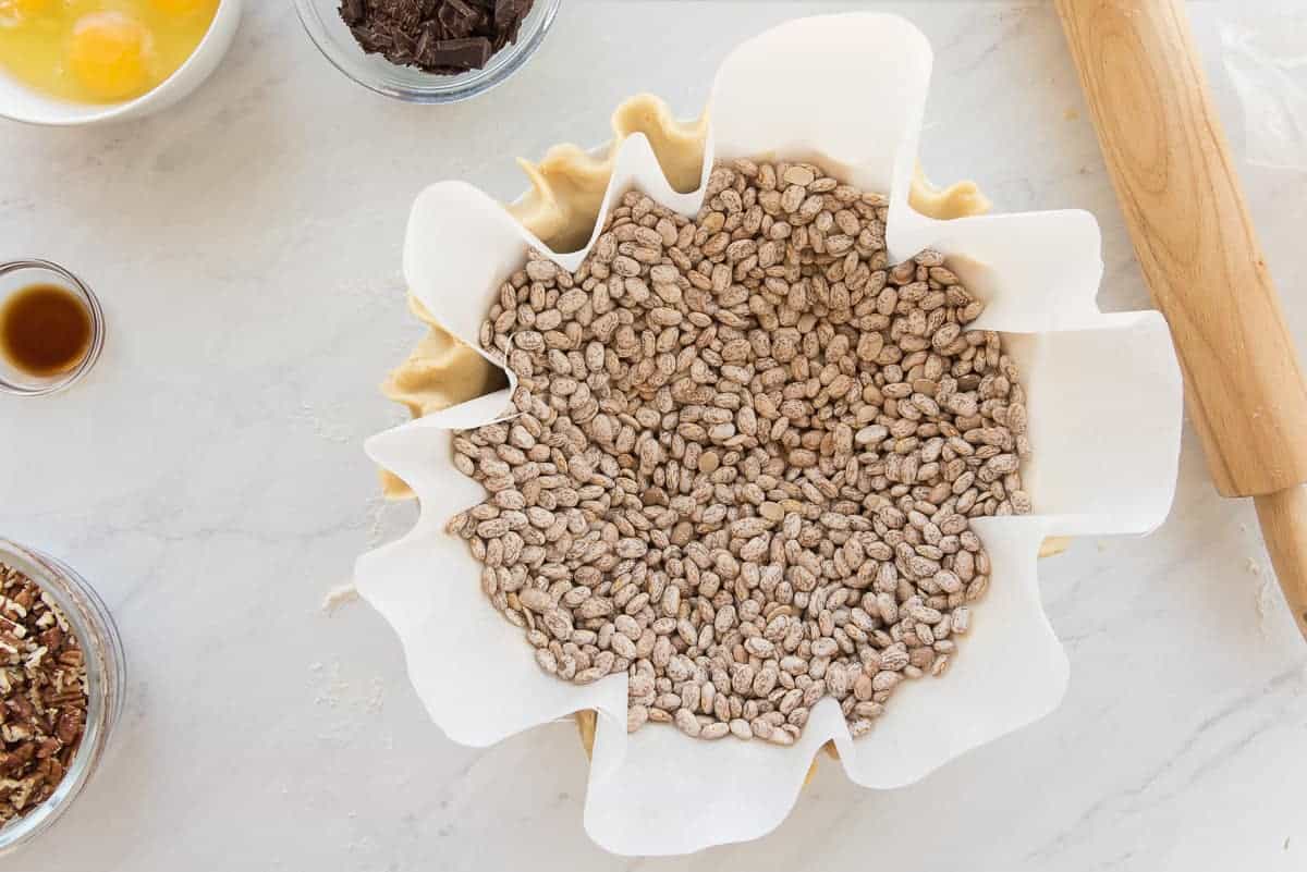 Dried beans are in a parchment paper liner before the pie dough is blind baked. A rolling pin, bowl of chocolate chunks, eggs, vanilla, and nuts surround the pie dish.