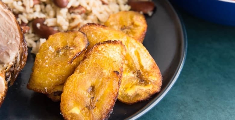 horizontal image of air fried sweet plantains on a dark plate next to rice and peas.