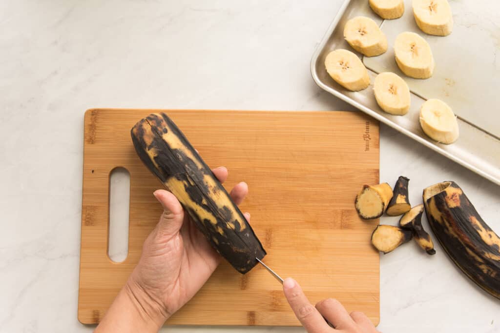 A hand makes a slit down the back of a black and yellow plantain with the tip of a knife.