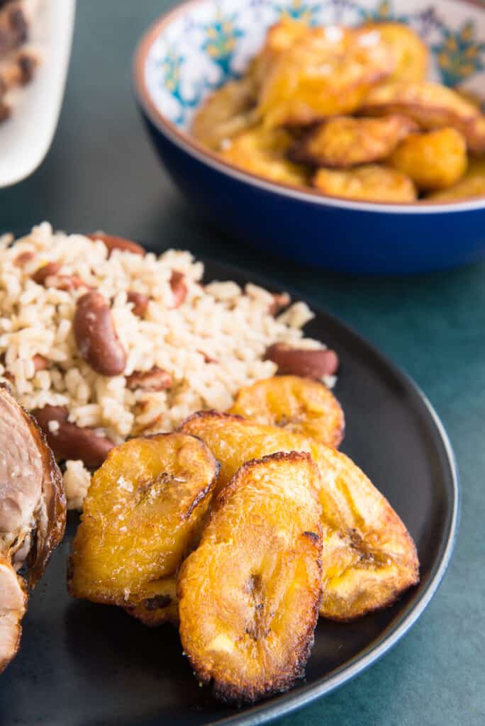 Lead portrait image of a pile of Air Fryer Plátanos Maduros (Sweet Plantains) on a dark blue plate next to a mound of Jamaican rice and peas.