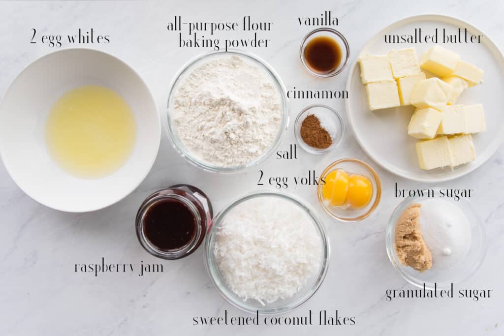 The ingredients for Coconut Raspberry Thumbprint Cookies: egg whites, all-purpose flour, baking powder, vanilla, cinnamon, unsalted butter, salt, egg yolks, brown sugar, granulated sugar, sweetened coconut flakes, and raspberry jam.