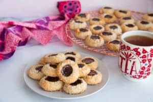 Horizontal image of a light grey plate of Coconut Raspberry Thumbprint Cookies next to a mug with the word TEA written on it in red