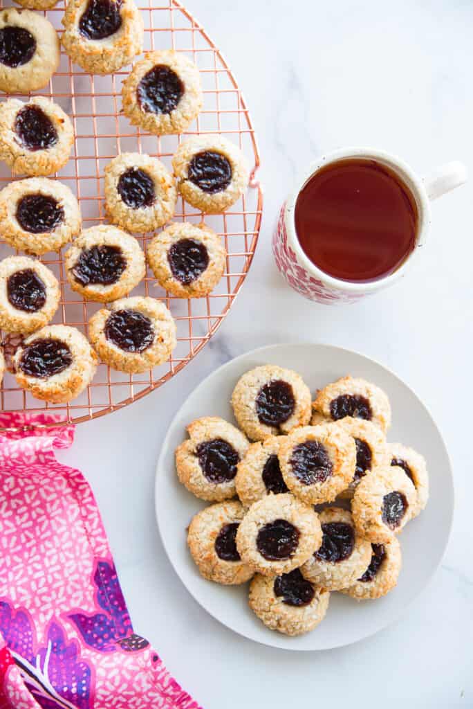 Overhead portrait image of a light grey plate of Coconut Raspberry Thumbprint Cookies next to a copper cooling rack with the rest of the cookies on it.