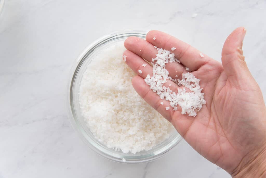 A hand lifts some of the coconut flakes from a clear glass bowl to show size.