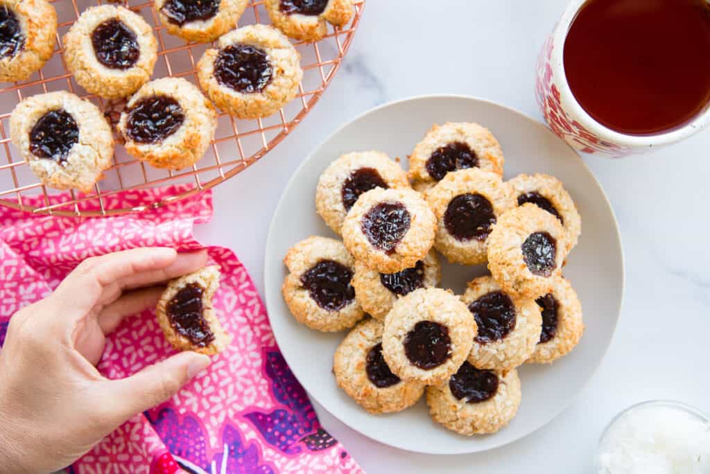A hand holds a Coconut Raspberry Thumbprint Cookies that has a bite taken from it. The light grey plate of cookies is next to a cream-colored mug of tea.