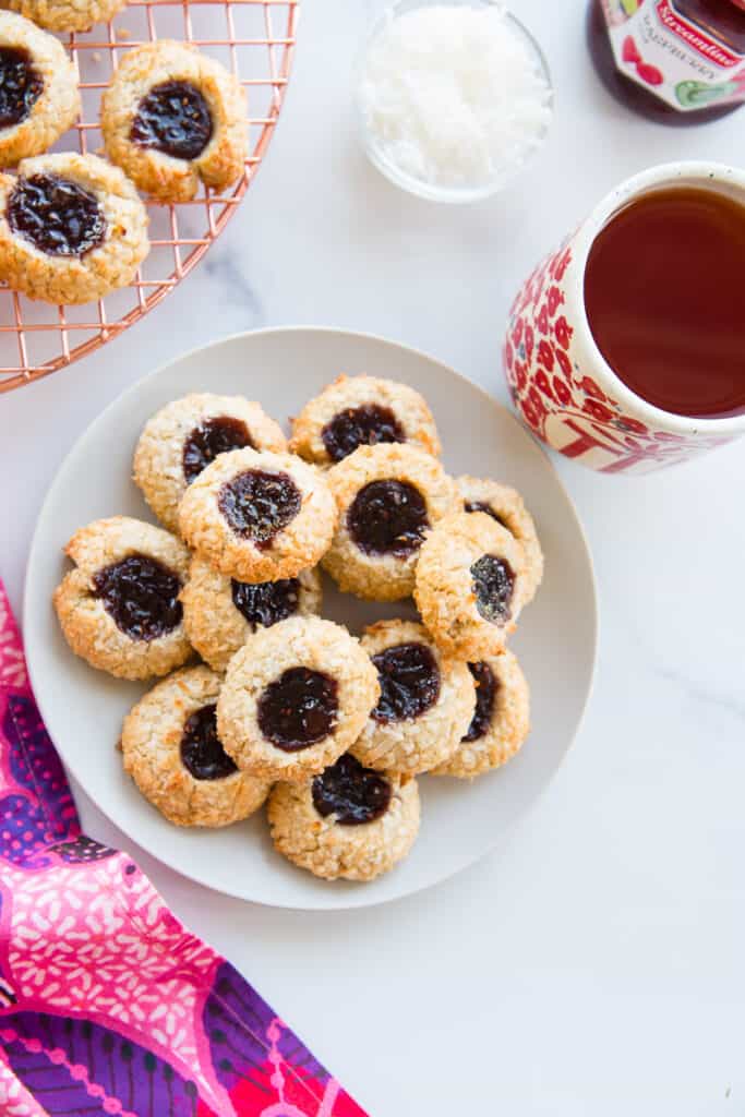 Overhead portrait image of a light grey plate of Coconut Raspberry Thumbprint Cookies next to a cream mug of tea and a copper cooling rack with more cookies on it.