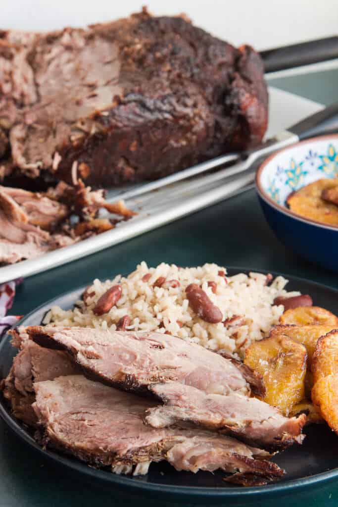 Portrait image of a dark plate with sliced Jerk Pernil, sweet plantains, and rice and peas in front of a platter with the pernil on it .