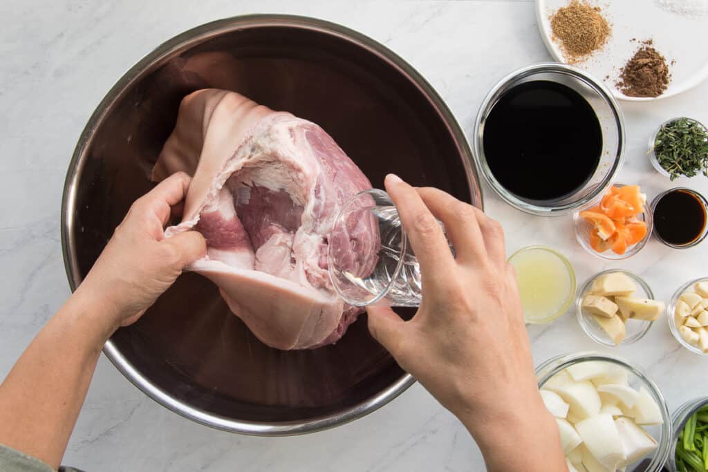 White vinegar is poured between the skin and meat of the pork shoulder which is in a metal bowl next to the rest of the jerk marinade ingredients.
