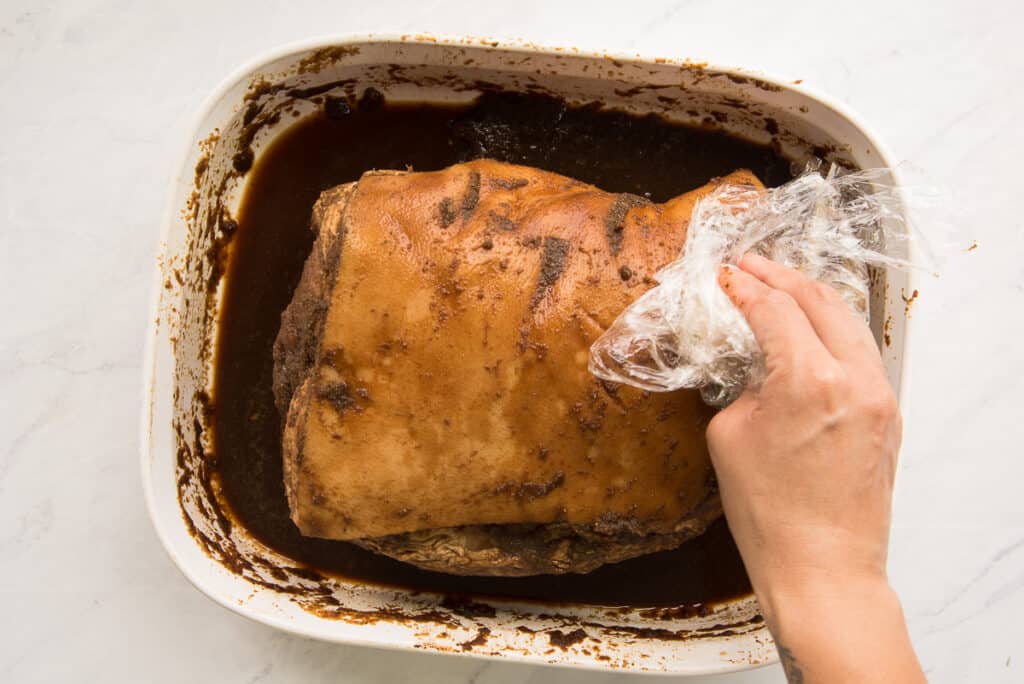 A hand uses the plastic wrap to wipe the jerk marinade off the surface of the skin.