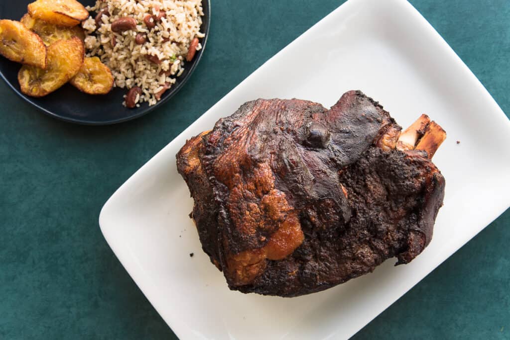 The roasted pork shoulder on a white platter next to a plate with rice and peas and sweet plantains.