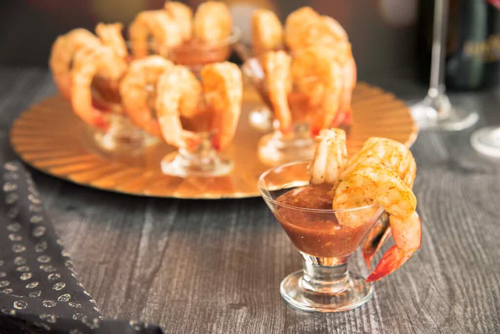 Horizontal image of a small appetizer glass with cocktail sauce and three roasted shrimp hanging off the right side of the rim.