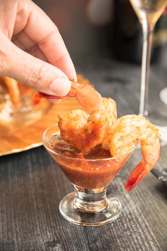 A hand dips a Roasted Shrimp into the homemade cocktail sauce in a small appetizer glass