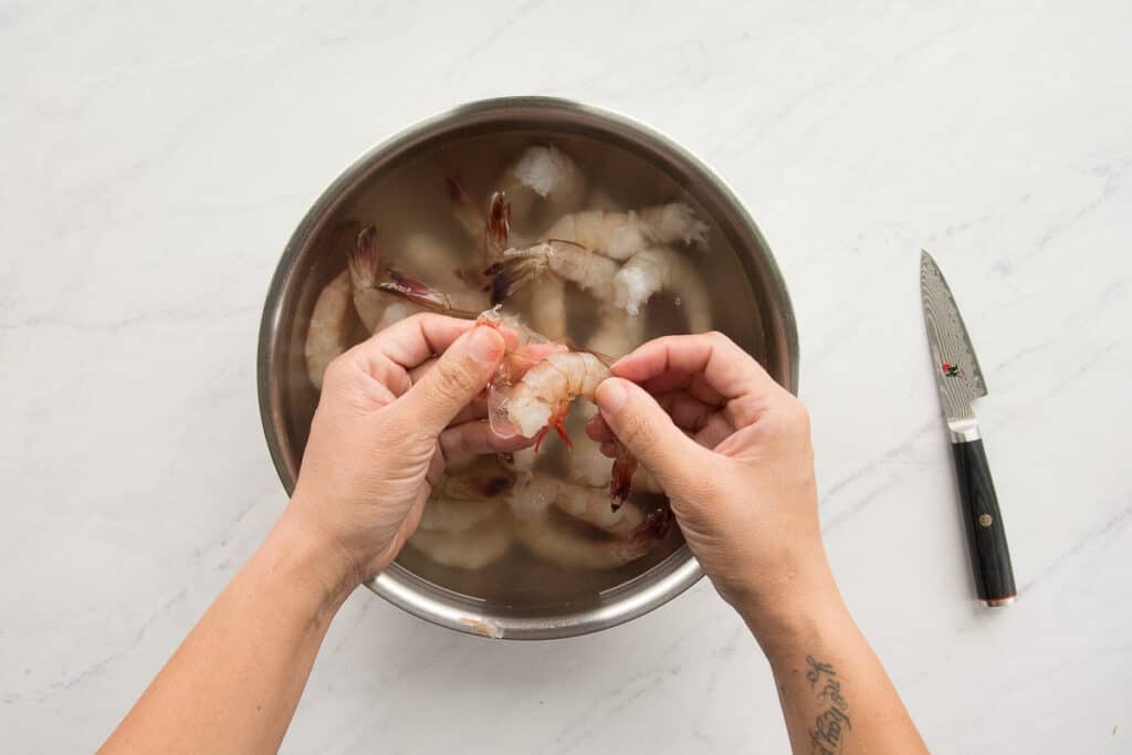 Hands removing the shell from a shrimp over a bowlful of shrimp in water.