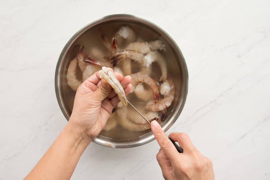 A hand holds a shrimp while using a paring knife to cut it down the back to devein it.