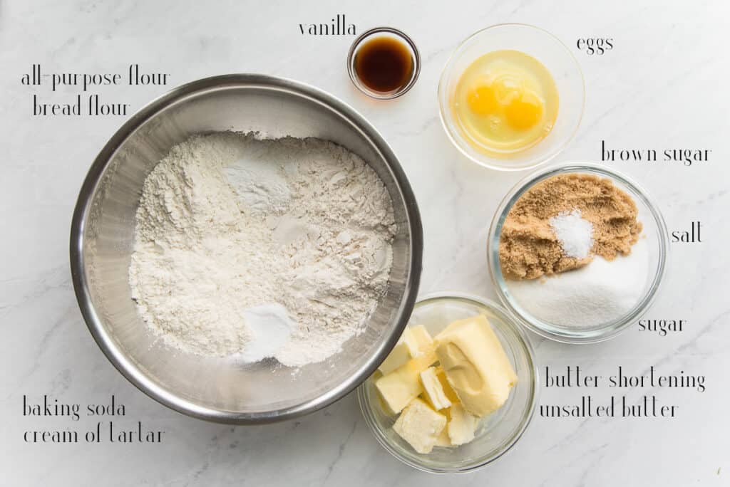 Ingredients to make the cookies on a white countertop: flours, leavening agents, salt, cream of tartar, vanilla, eggs, brown and white sugar, shortening, and unsalted butter.