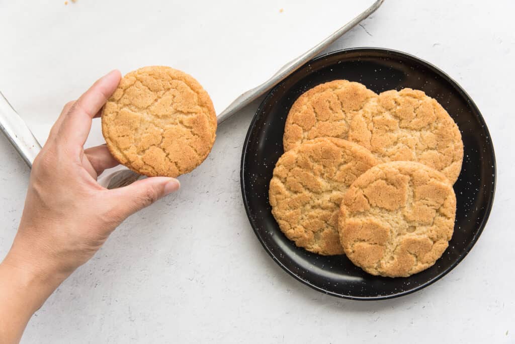 A hand lifts a snickerdoodle cookie from a sheetpan to put it on a black plate of cookies.