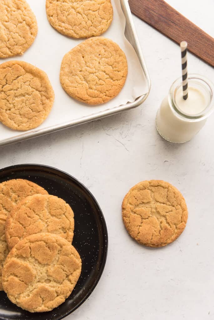 Lead image of a sheetpan of baked snickerdoodle cookies next to a bottle of milk and a single cookie. A plate of cookies is at bottom left.