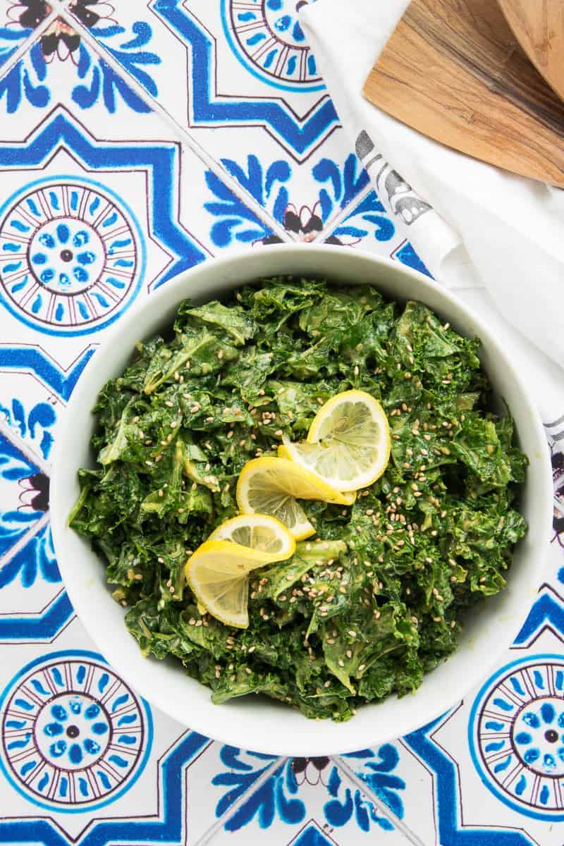 Overhead portrait image of a white bowl of Kale with Lemon-Tahini Dressing on a blue and white tiled surface.