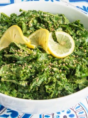 Preview image of a white bowl filled with Kale with Lemon-Tahini Dressing garnished with sesame seeds and a twisted lemon slice.