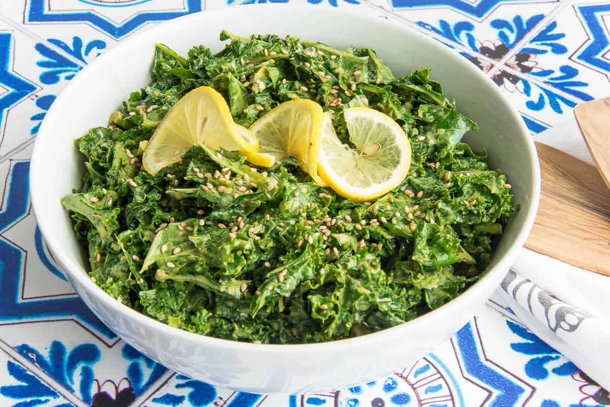 Preview image of a white bowl filled with Kale with Lemon-Tahini Dressing garnished with sesame seeds and a twisted lemon slice.