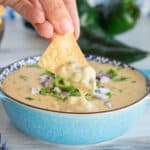 Social media image of a hand lifting a chip dipped in Roasted Poblano White Queso Dip from a blue bowl.