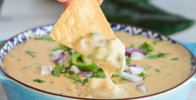 Social media image of a hand lifting a chip dipped in Roasted Poblano White Queso Dip from a blue bowl.