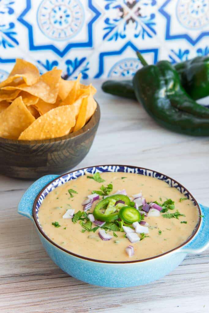 Lead image of a light blue bowl filled with Roasted Poblano White Queso Dip. Poblanos and a brown bowl of tortilla chips is in the background.