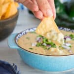 Preview image of a hand lifting a corn tortilla chip from a blue bowl filled with Roasted Poblano White Queso Dip