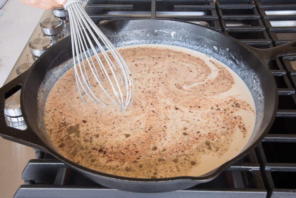 Spices are added to the thickened mixture with a grey whisk in a black cast iron skillet.