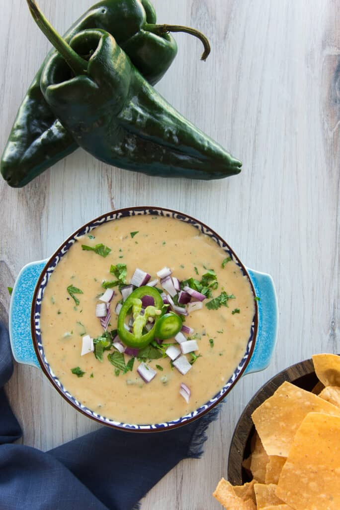 Overhead portrait image of the Roasted Poblano White Queso Dip in a blue bowl next to two poblano peppers and a bowl of tortilla chips.