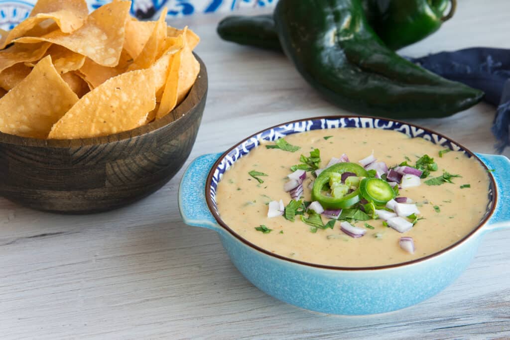 Horizontal image of a blue bowl of Roasted Poblano White Queso Dip garnished with cilantro, jalapeño slices, and chopped red onion. Poblano peppers and corn tortilla chips in a wooden bowl are in the background.