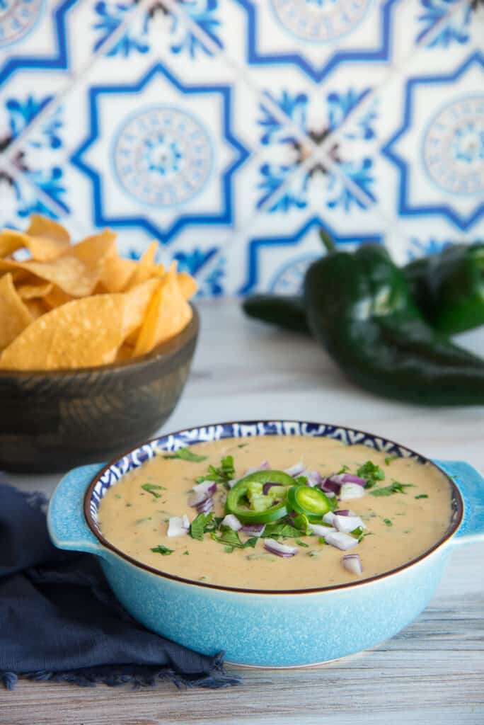 The Roasted Poblano White Queso Dip is in a light blue bowl, garnished with jalapeño slices, cilantro, and red onion. Poblano peppers and a brown bowl of corn tortilla chips is in the background.