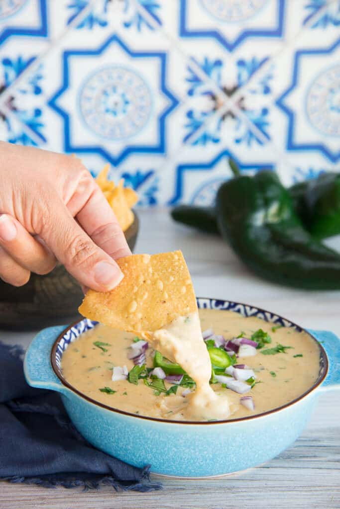 Portrait image of a hand dipping a tortilla chip into a bowl of Roasted Poblano White Queso Dip