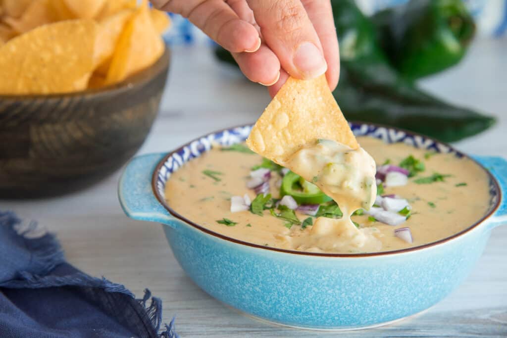 A horizontal image of a hand lifting a chip dipped into Roasted Poblano White Queso Dip that's in a blue bowl.