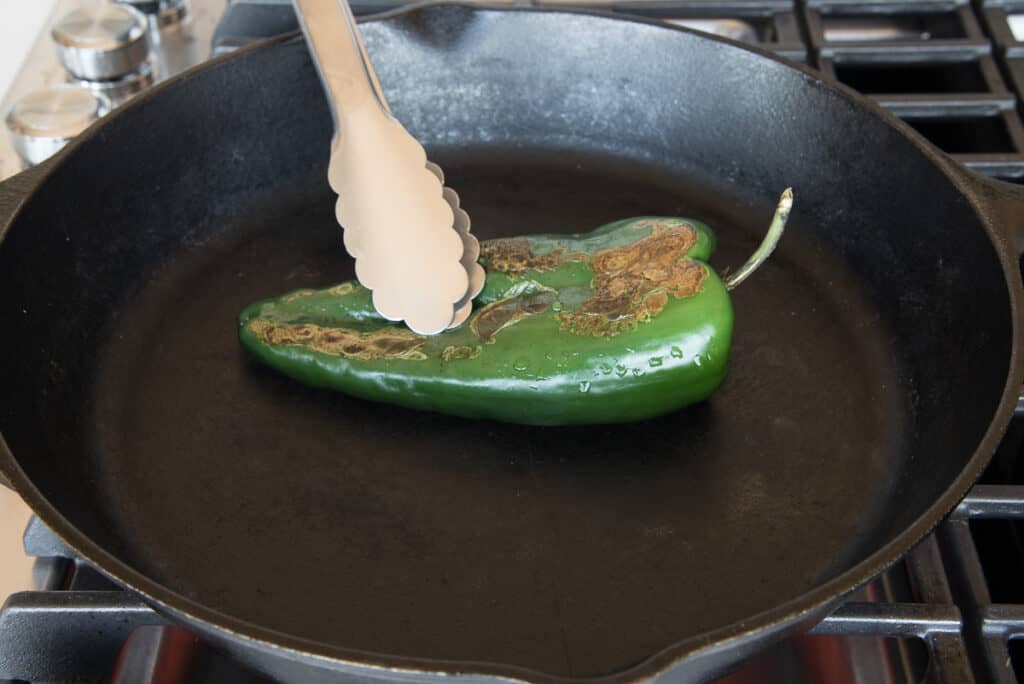 Tongs press down the green pepper, which is blistered on top, in a black cast iron skillet.