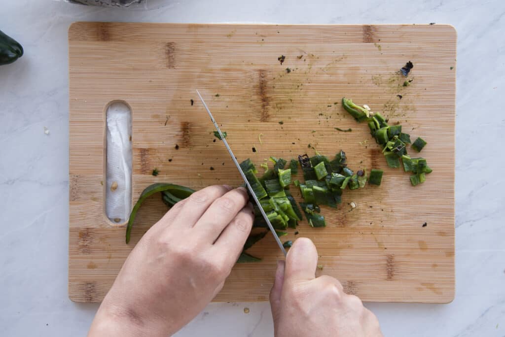 Hands hold a knife and the peeled pepper to dice it on a wooden cutting board.