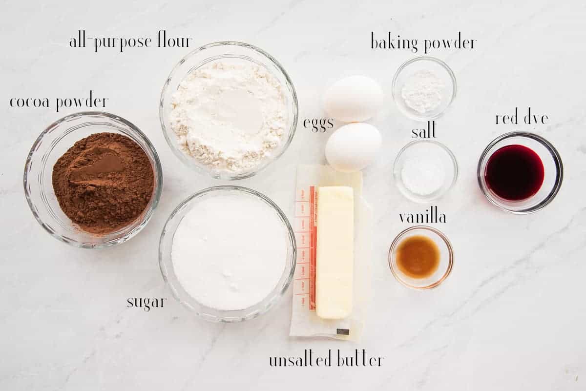 Ingredients to make the red velvet brownie shown on a countertop: cocoa powder, all-purpose flour, sugar, eggs, baking powder, salt, vanilla, and red dye.