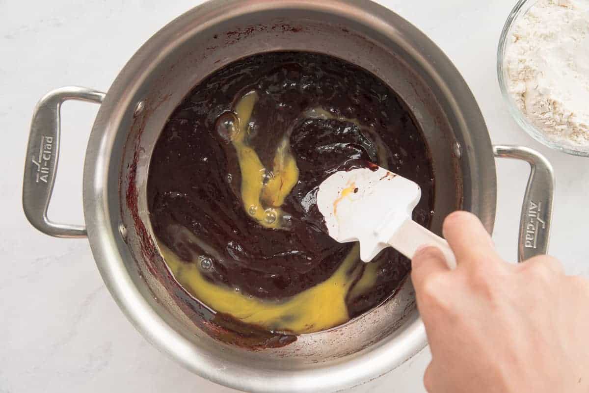 Eggs are added to the cocoa mixture in a silver pot with a white rubber spatula.