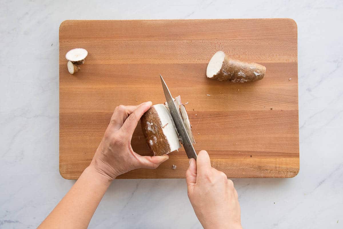 A knife is used to remove the waxy peel from a section of yuca on a wooden cutting board.
