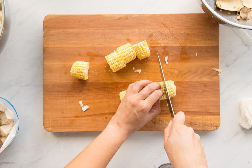 a knife is used to cut corn cobs into 2-inch wide slices on a wooden cutting board.