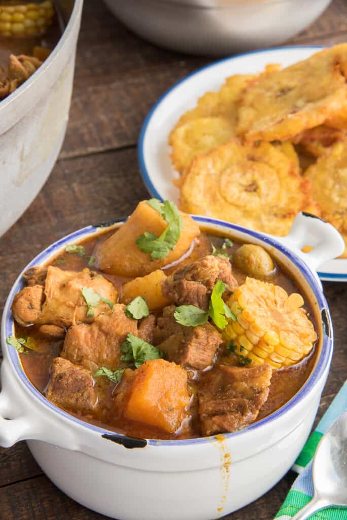 Close-up portrait of a bowl of Sancocho in front of a plate of tostones.